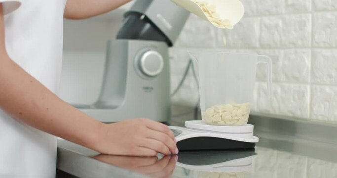Close up image of young professional pastry chef, weighing ingredients for ganache or cream, putting white chocolate drops in measuring cup. Confectionery shop, baking concept.