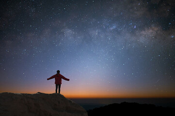 Silhouette of young traveler and backpacker watched the star and milky way alone on top of the mountain. He enjoyed traveling and was successful when he reached the summit. - obrazy, fototapety, plakaty