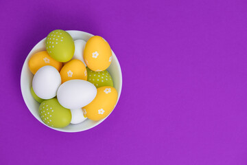 Colored eggs in a white plate on a purple background. Easter eggs. Copy space. Spring mood with yellow, white and green chicken eggs. 