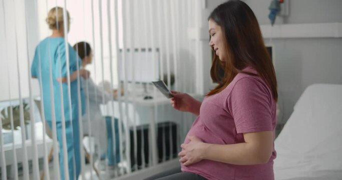 Young pregnant woman looking at ultrasound image sitting at bed in hospital