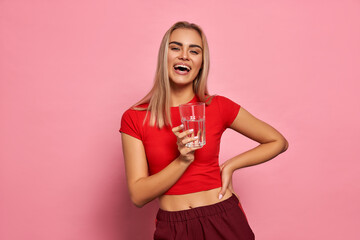 Smiling blonde woman in sport suit holding a glass of water isolated on pastel background