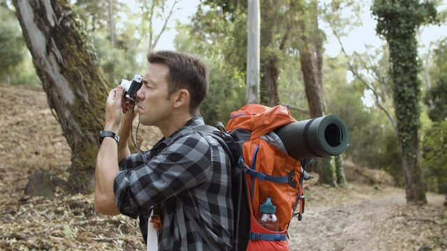 Active tourist guy with photo camera taking pictures of forest landscape, while walking on path, enjoying outdoor leisure time and healthy recreation. Adventure travel concept