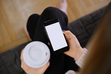 Back view close up of a woman hand holding and using smart phone with white blank empty screen sitting on a bed at home