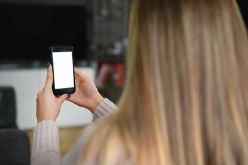 Back view close up of a woman hand holding and using smart phone with white blank empty screen sitting on a bed at home