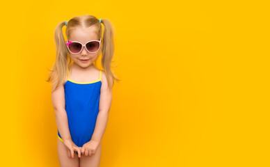 Mockup with little cute baby posing in a bathing suit on an isolated yellow background. Beautiful cute girl in a blue swimsuit with ponytails and sunglasses in a model pose. Banner. Place for text