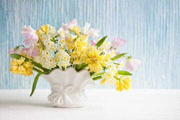 Spring bouquet of yellow hyacinths, daffodils and pink sweet peas in a vase on a table, blue background wall. Closeup, blur, soft focus.