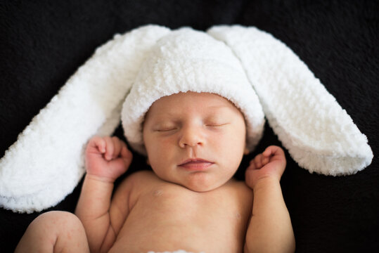 Naked newborn baby in bunny knitted white costume on black background. Close-up.