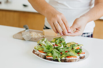 Young man making toasts with avocado, tomatoes, arugula, cheese in modern kitchen, putting parmesan