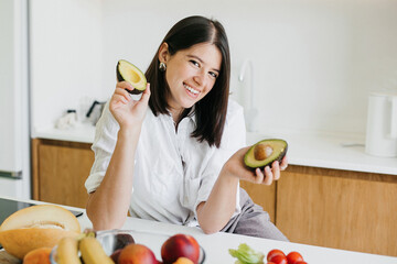 Young happy woman holding halves of ripe avocado and smiling in modern kitchen. Healthy eating