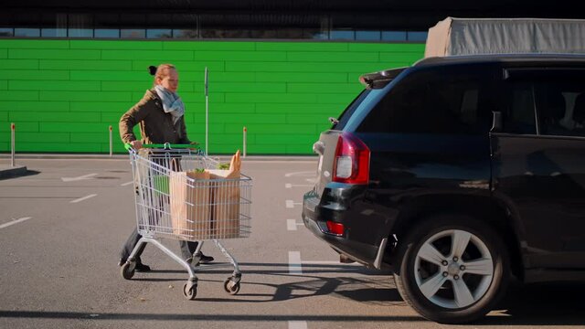 adult caucasian woman going with shopping cart to the car parking near shopping mall or store