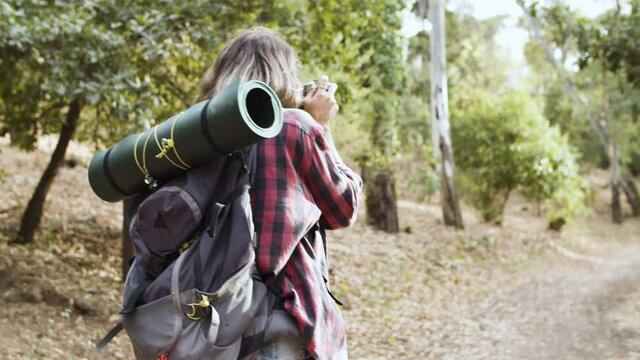 Young female backpacker with photo camera taking pictures of forest landscape while walking on path, enjoying outdoor leisure time and healthy recreation. Adventure travel concept