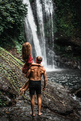 A man of athletic build holds a beautiful girl in his arms at the waterfall. Couple at the waterfall, rear view. Honeymoon trip. Happy couple on vacation in Bali. A couple in love travels the world.