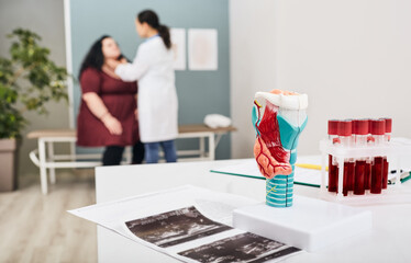 Thyroid anatomical model and thyroid hormone blood test tubes, close-up. Doctor probes the neck of a fat woman in background