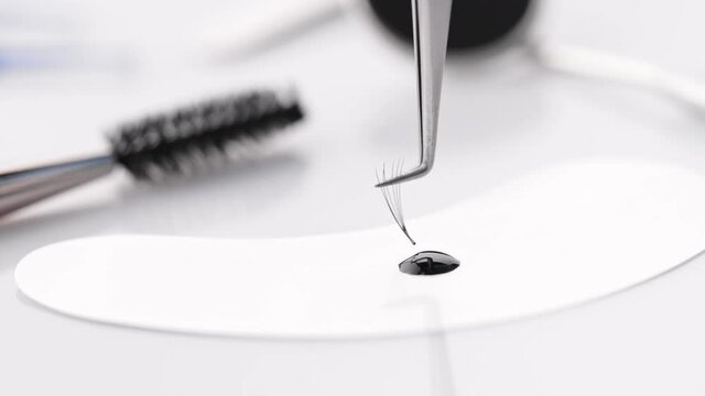 Accessories for eyelash extensions. Beauty master holding curve tweezers with false eyelash, dip it into drop of eyelash glue. Tools for eyelashes extension procedure.