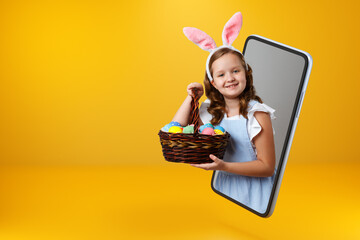 A little girl in bunny ears holds a basket with colorful Easter eggs and looks out of a smartphone. Child online on a mobile phone screen. Copy space.