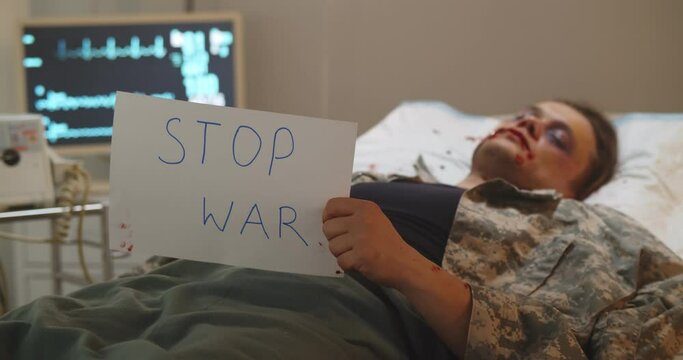 Wounded and weak soldier lying in hospital bed and holding stop war poster