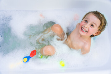 Little boy playing with toy by taking  bath in bathtub. Smiling toddler  sitting in bathtub with lot of fluffy soap bubble and smiling. Daily routine with children.