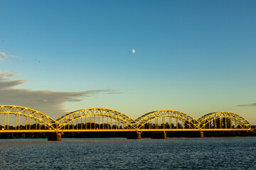 Riga´s railway Iron Bridge over Daugava River during sunset what makes it yellow, river water dark blue, half-moon is shining, black birds are flying in gradient blue sky 