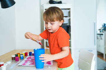 Little boy sitting at the desk and drawing colorful picture with paint and brushes. Child education at home during self isolation and lockdown. Concept of art and creativity of children.