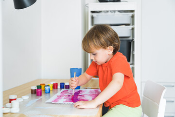 Little boy sitting at the desk and drawing colorful picture with paint and brushes. Child education at home during self isolation and lockdown. Toddler does not know how to hold a brush correctly.