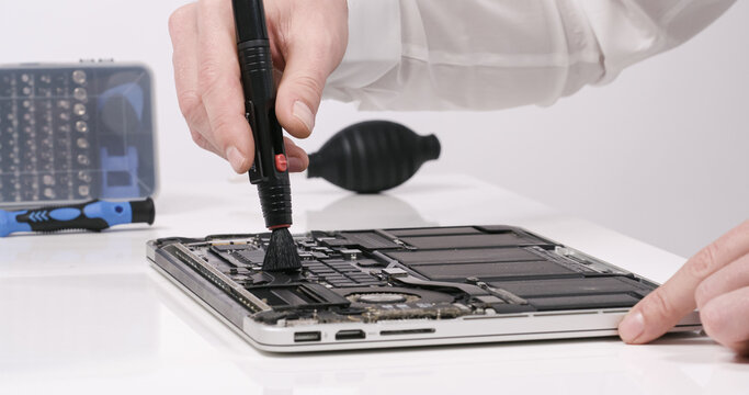 Service worker repairing cleaning dust laptop motherboard circuit board cooler with brush in white walled lab 