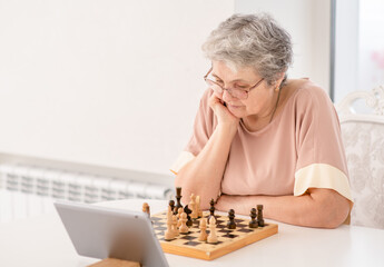 Pensive senior woman uses tablet computer to play chess with her friends during quarantine...
