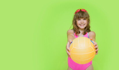 A beautiful child in a pink swimsuit shows a beach volleyball ball to the camera. Plortret of a cute little girl with swimming goggles on a green background. The concept of summer holidays.