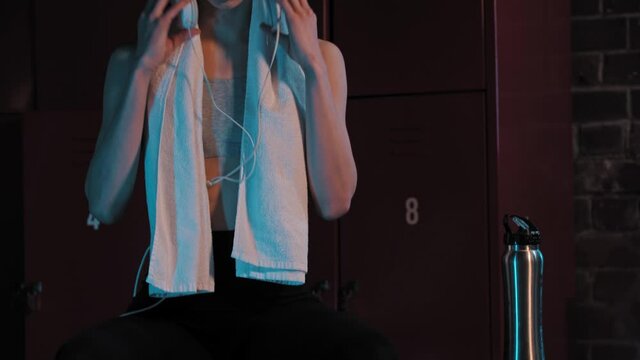 A young pretty woman sitting in locker room with a towel and headphones on her neck