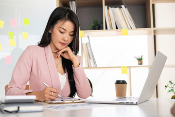 Positive Asian female finance is writing some notes for tomorrow's meeting with the foreign company using phone and notebook. A beautiful financial manager dressed in a pink suit in the office.