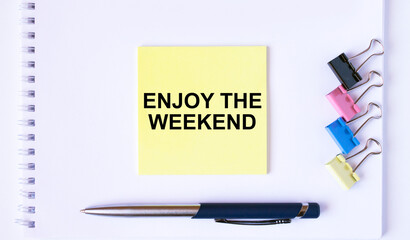 Yellow sticker with text Enjoy The Weekend lying on a white Notepad with a pen and paper clips