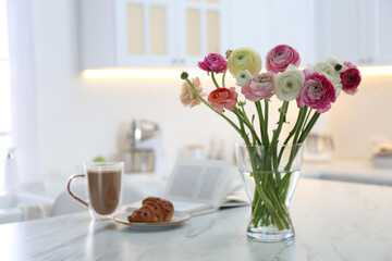 Beautiful fresh ranunculus flowers near cup of coffee, croissant and book on table in kitchen