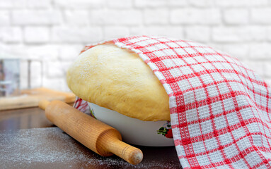Raw yeast dough covered with a towel in a bowl on the floured kitchen table, recipe idea. Concept home baking or making dough.