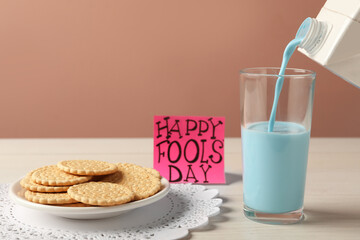 Pouring light blue milk into glass, cookies and words Happy Fool's Day on table