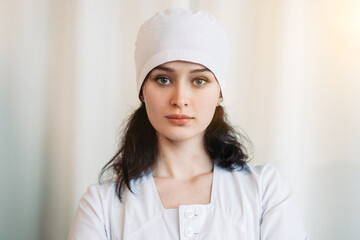 Nurse young medic standing near white wall and looking at the camera. Female doctor dressed in medical gown and surgical hat. Image with copy space. Pandemic virus coronavirus concept
