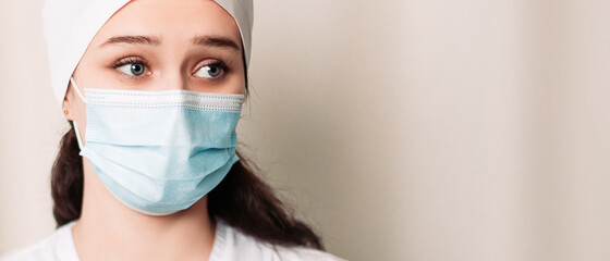 Nurse young medic standing near white wall and looking at the camera. Female doctor dressed in medical gown and surgical hat. Pandemic virus coronavirus concept, disposable face mask, copy space.
