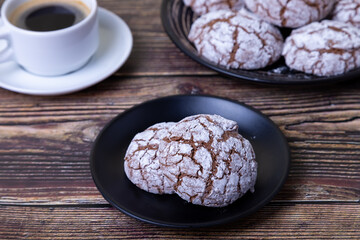 Chocolate cookies with cracks, strewed with icing sugar. Homemade baking, wooden background. Background: a cup of coffee and a plate of cookies. Selective focus, close-up