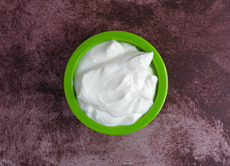 Overhead view of a small green bowl filled with plain Greek yogurt on a marron tabletop illuminated with natural light.