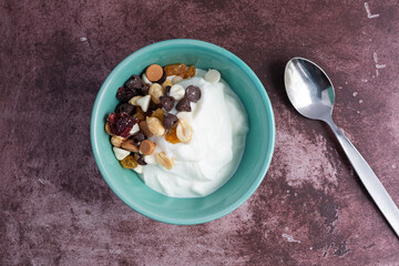 Overhead view of a bowl with plain Greek yogurt and a candy trail mix on a marron tabletop with a spoon to the side illuminated with natural light.