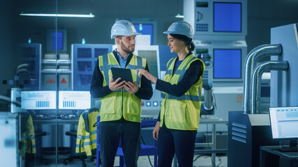 Modern Factory: Female and Male Engineers Wearing Safety Jackets, Hardhats Standing in Industrial Workshop, Talking and Using Tablet Computer. Facility with CNC Machinery, Robot Arm Production Line