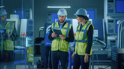 Modern Factory: Female and Male Engineers Wearing Safety Jackets, Hardhats Standing in Industrial Workshop, Talking and Using Tablet Computer. Facility with CNC Machinery, Robot Arm Production Line