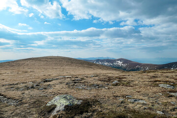 A panoramic view on an Alpine chain in Austria. The vast pasture has golden colors. There are other mountain chains in the back. A narrow pathway leading to the top. Overcast. Serenity and calmness