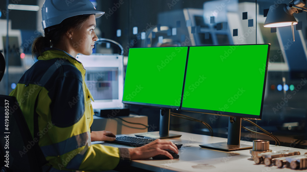Poster young and confident industrial female engineer in safety vest and hardhat is working on computer wit