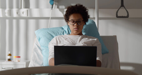 Young african female patient working with laptop in bed in hospital