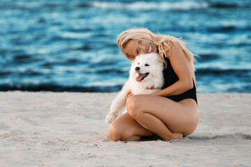 Young beautiful blonde girl playing with white samoyed puppy on the windy beach in the black bikini