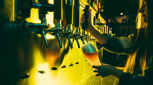 Young woman barman pouring a cold lager beer from tap to glass in neon light.