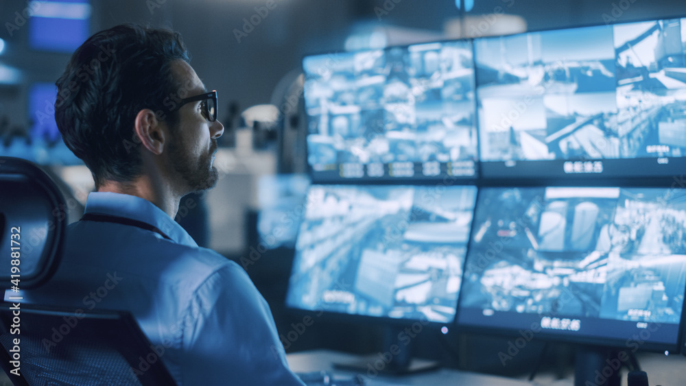 Poster Industry 4.0 Modern Factory: Portrait Shot of a Security Operator Controls Proper Functioning of Workshop Production Line, Uses Computer with Screens Showing Surveillance Feed. High-Tech Security
