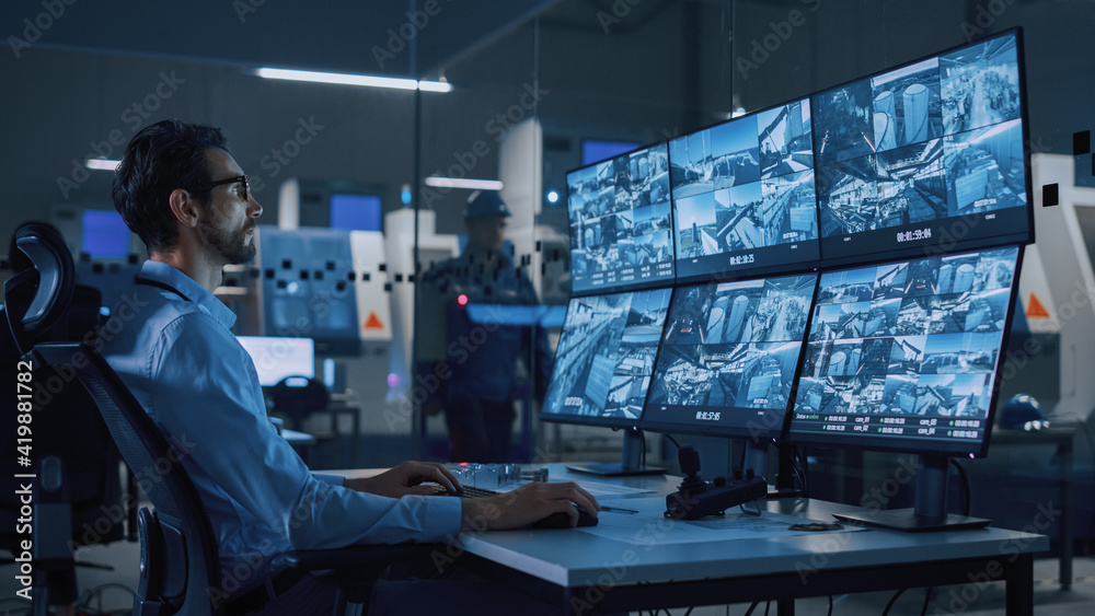 Fototapete Industry 4.0 Modern Factory: Security Operator Controls Proper Functioning of Workshop Production Line, Uses Computer with Screens Showing Surveillance Camera Feed. High-Tech Security