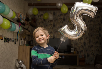 handsome cute boy in a sweater celebrates his birthday, holds a silver number 5 in his hands. The room is decorated with green balloons in the background. Celebrating a joyful event