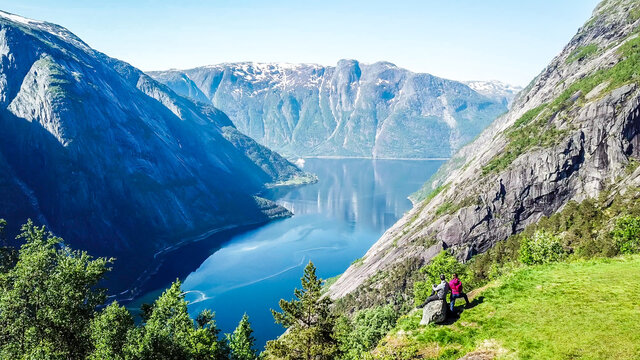 A couple standing on the meadow with a majestic view on Eidfjord from Kjeasen, Norway. Slopes of the mountains are overgrown with lush green grass. Water has dark blue color. Sunny and clear day