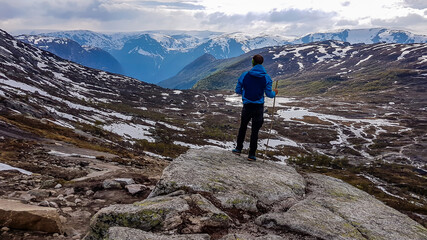 Fototapeta premium A young man wearing blue jacket stands on a pile of rocks and admires the view in front of him. He supports himself on a wooden stick. Endless mountain ranges partially covered with snow. Big overcast